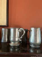 Four pewter mugs and tankards arranged on surface, showing varying shapes and sizes including decorative handle and inscriptions.