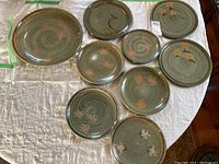 View of nine ceramic plates and a large platter arranged on fabric. Green glaze with spiral texturing and orange floral designs visible.