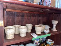 Six ceramic cups, goblet, and small bowls arranged on a wooden shelf with some other pottery below.