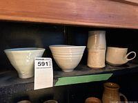 Front view of 2 white bowls, 1 tall beige cup, and 1 small cream cup displayed on a shelf.