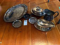 Full view of silver plated platter, sauce boat with under plate, water jug, covered vegetable server, small plate, divided condiment dish, and small round plate on wooden floor.