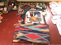 View of assorted souvenir textile lot on floor with rugs, belts, doll, bag, and coasters