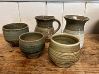 Five pottery pieces including a mug, tall cup, and three smaller cups in green and earth tones on wooden surface