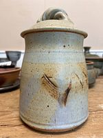 Front view of the signed pottery vessel showing earthy glaze and lid with loop handle.