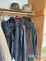 Side view of four coats on hangers showing black leather coat, brown suede coat, and two wool coats, with felt and lamb's fur hats on shelf above