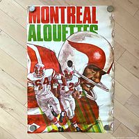 Full view of vintage Montreal Alouettes football poster showing three players in white uniforms with red and green helmet and jersey detail. The poster is laid flat on a wooden floor and secured by four large coins at the corners for display purposes.