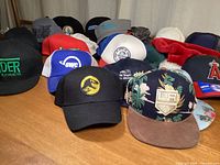 Photo showing a collection of caps with various colors and logos including Vader Extracts, dinosaur logo, floral pattern cap, and OWC trucker hats on a wooden surface.
