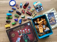 All items displayed on wooden floor, multiple vintage die-cast cars including Hot Wheels and Matchbox, wooden mecha model parts in blue plastic box with clear lid, and Hovering Ball Shooting Game box with visible ball shooter and targets.