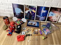Photo showing various Disney and Marvel collectible toys, prints, keychains, and memorabilia arranged on the floor including lenticular Lion King frame, Cinderella prints, Spider-Man figures, buttons, and Astro Blaster toy.