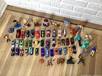 Full layout of Happy Meal toys and assorted small toy cars on wooden floor against white brick wall.