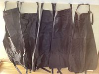 Lot of five black aprons laid out flat showing full view of pieces with neck straps and waist ties.