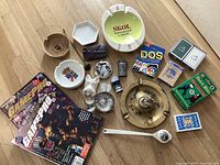 Overview photo showing all lot items on wooden floor including GamePro magazines, various ashtrays, wine tasting dish set, smoking accessories, and decks of playing cards.