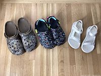 Top view of three pairs of shoes: brown camouflage Crocs, black splatter Crocs with colorful paint splatters, and white Tevas sandals.