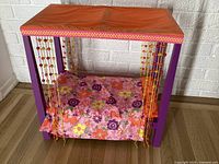 Front view of purple wooden doll bed with orange fabric canopy top, floral bed cover, and beaded curtains hanging on each side.