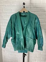 Front view of teal leather jacket hanging on wooden hanger, showing patchwork leather design and billowy sleeves.