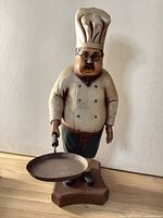 Front view of vintage chef figurine showing detailed face, chef hat, double-breasted chef coat, apron, and frying pan held in right hand.
