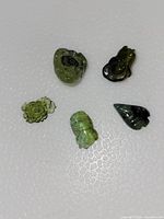 Top-down photo showing five green tourmaline carvings on a textured light surface: three flowers, one fish, one leaf.