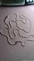 Two silver snake chains laid out on a surface showing full length and clasp.