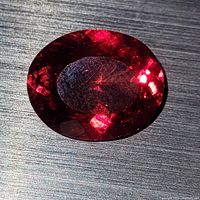 Close-up photo of oval-shaped deep red faceted almandine garnet on a dark textured surface showing internal reflections and facet details.