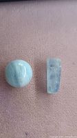 Photo showing raw aquamarine crystal and polished marble sphere placed on a fabric surface, demonstrating size and details