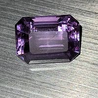 Top-down clear photo of emerald cut purple sapphire on a textured surface showing facets and internal reflections.