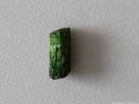 Raw green diopside crystal on white textured background showing its natural prismatic structure and shine.