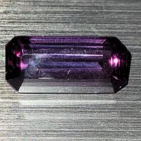 Top view of emerald cut sapphire in indoor lighting showing deep purple color, visible faceting and internal characteristics.