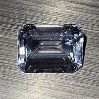 Close-up image of a rectangular aquamarine stone with emerald cut facets, showing light blue color and clarity against a textured background.