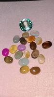 Photo of a large round faceted green man-made tourmaline and 28 oval polished cabochons in varied colors including green, yellow, brown, pink, purple, and light blue on a soft pink background.