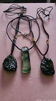Three necklaces laid out: two with rough textured dark green moldavite pendants and one with smooth green art glass pendant. All on black adjustable cords with bead accents.