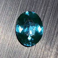 Close-up of oval neon blue-green apatite gemstone showing surface facets and reflective qualities.