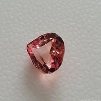 Close-up photo of a pear-shaped pink/peach padparadscha sapphire gemstone on a white textured background.
