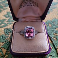 Front view of the pink sapphire ring in a vintage style ring box, showing main cushion-cut pink sapphire stone and white topaz accents on silver band.
