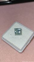 Front view of princess cut diamond on white foam pad in plastic display box, showing blue-green tint