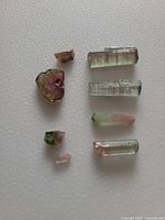 Top view of seven raw watermelon tourmaline stones laid out on white surface showing varied shapes and colors from pink center to green edges.