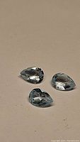 Three pear cut sky blue topaz gemstones positioned on a neutral beige surface showing their shape, color, and clarity.