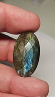 Photograph of the large oval labradorite gemstone held against fingers, showing blue and golden flash with cabochon cut surface.