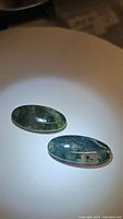 Two oval cabochon moss agate stones on a white surface under light showing moss-like green inclusions.