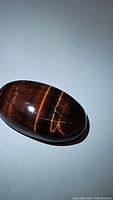Close-up image of a polished oval cabochon red tigerseye gemstone showing its characteristic chatoyancy and rich reddish-brown tones.