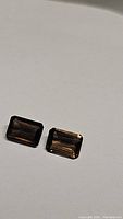 Two natural smokey quartz gemstones shown side by side on a light neutral background, highlighting their emerald cut and dark smoky color.