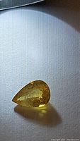 Photo of a pear-shaped natural yellow sapphire with visible inclusions, warm golden yellow color, sitting on light textile background.