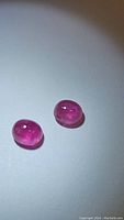 Photo showing two deep pink oval cabochon sapphires placed on a white surface under bright light. Gems show translucency and internal inclusions.