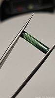 Long octagon cut green tourmaline gemstone held with tweezers showing the length and color from the side profile