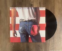Vinyl LP of Bruce Springsteen's 'Born In The USA' album partially pulled out of original album cover, showing cover artwork of man with jeans and red cap against American flag background.