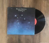 Photo of Willie Nelson Stardust LP vinyl record partially slid out of its album sleeve, showing the red label on the vinyl disc and starry sky album cover.