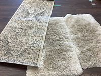 Photo showing the long patterned runner rug beside two light cream shaggy bath mats on a dark floor, illustrating size and texture.