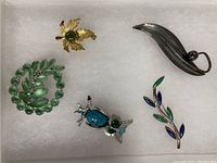 Five brooches displayed on white cushioning, showing variety including green rhinestone circular brooch, gold-tone maple leaf, silver-tone curved leaf with pearl, turquoise owl, and blue-green leaf brooch.
