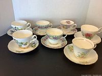 Seven vintage porcelain cups on matching saucers, each with distinct floral designs and gold trims, displayed on white background.