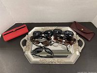 Five pairs of sunglasses and one pair of metal framed glasses on decorative mirrored tray with two hard cases on sides.