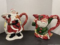 Two ceramic Christmas pitchers side by side. Left is Santa Claus with candy cane handle, right is elf pitcher with red hat lid and decorative holiday scenes.